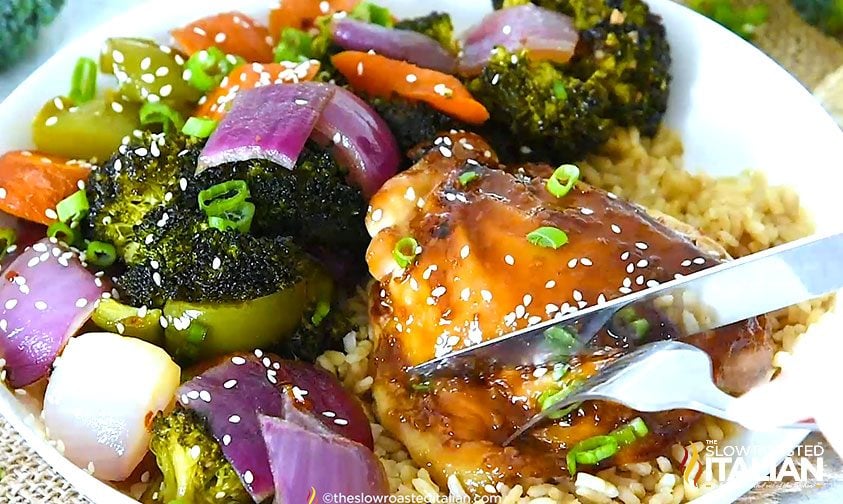 cutting into baked teriyaki chicken thigh on plate with rice and veggies