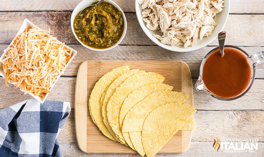 overhead: corn tortillas next to bowls of spicy red sauce, diced chiles, shredded cheese and shredded chicken