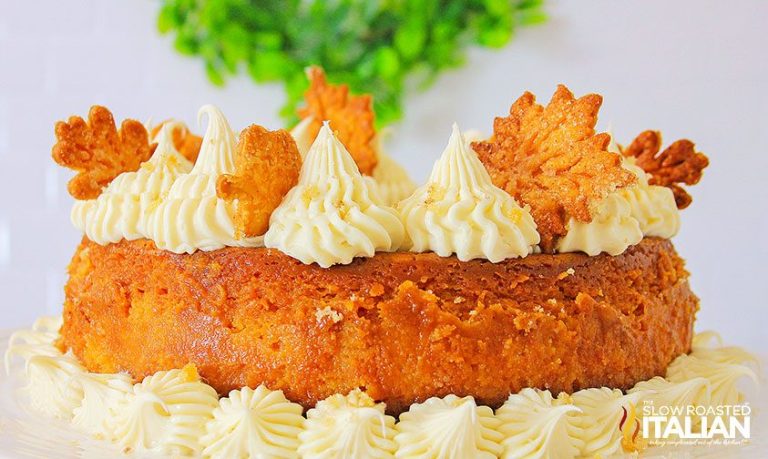 A pumpkin cake topped with swirls of white frosting and decorated with leaf-shaped pastry accents. The cake is set against a backdrop of green foliage, creating an autumnal theme.