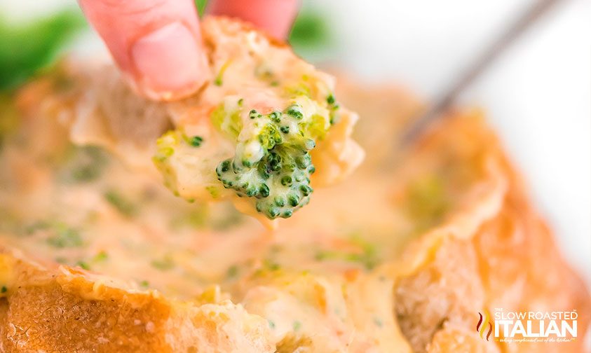 hand dipping piece of bread bowl into broccoli cheese soup