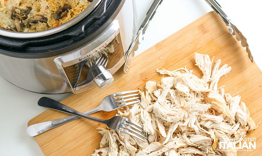 shredding chicken breast with forks on cutting board in front of pressure cooker