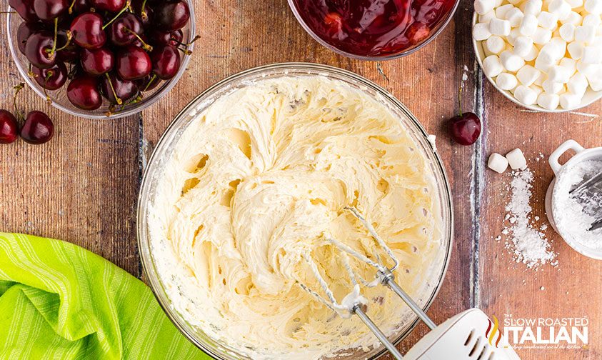beating cream cheese with electric mixer in a clear glass bowl