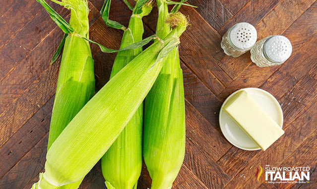 four ears of corn in husk next to plate of butter, salt and pepper