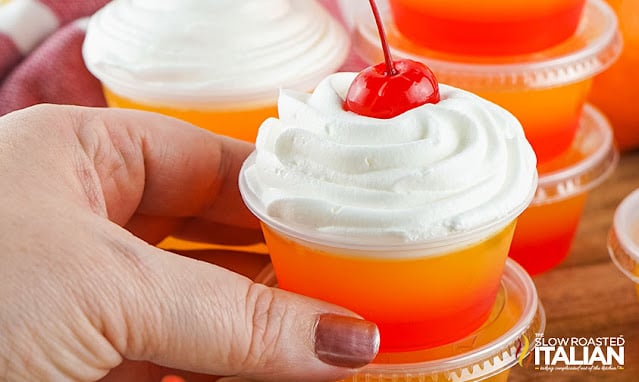 woman's hand holding tequila jello shot topped with whipped cream and a cherry