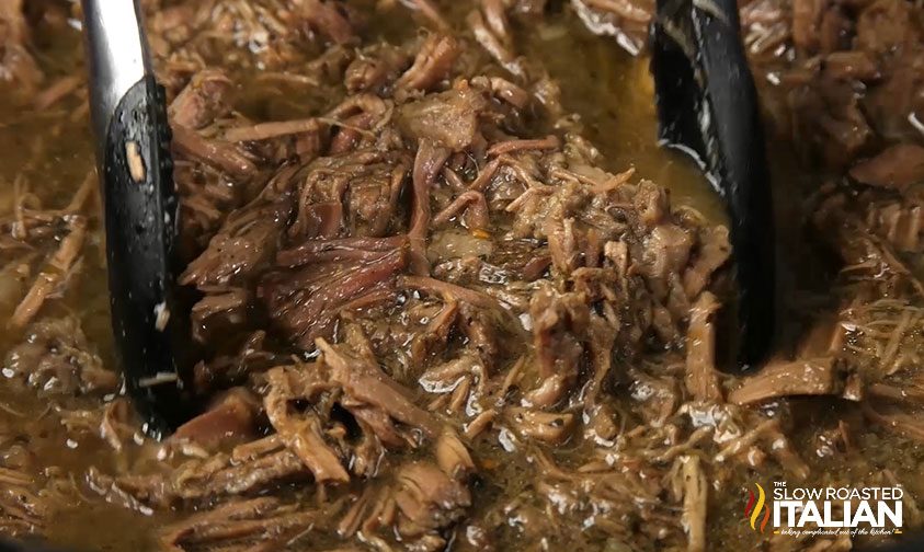 close up: tongs grabbing shredded italian beef in slow cooker