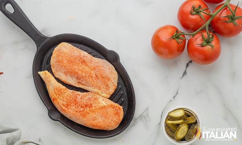 seasoned chicken on grill pan next to tomatoes and pickle slices