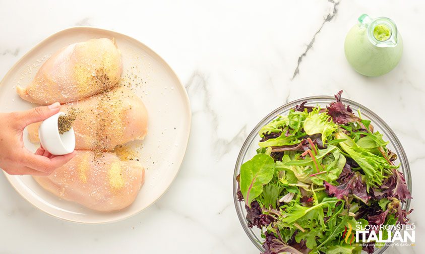 seasoning chicken breasts next to a bowl of spring mix and dressing