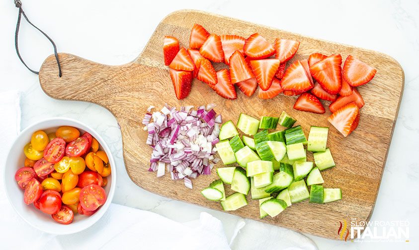 chopped strawberries, red onion, and zucchini on a cutting board with bowl of halved grape tomatoes