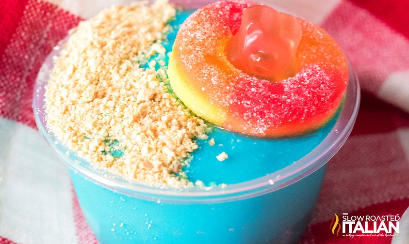 blue pudding shot topped with graham cracker crumbs and a gummy bear in a peach gummy ring