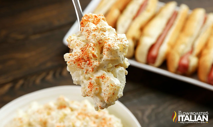 close up: spoonful of southern potato salad with tray of hot dogs in the background