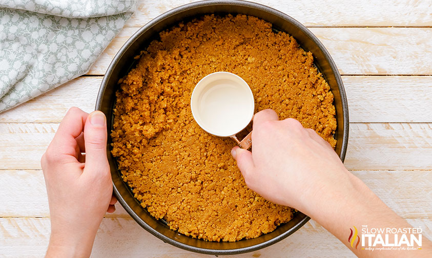 pressing graham cracker crust into springform pan with measuring spoon