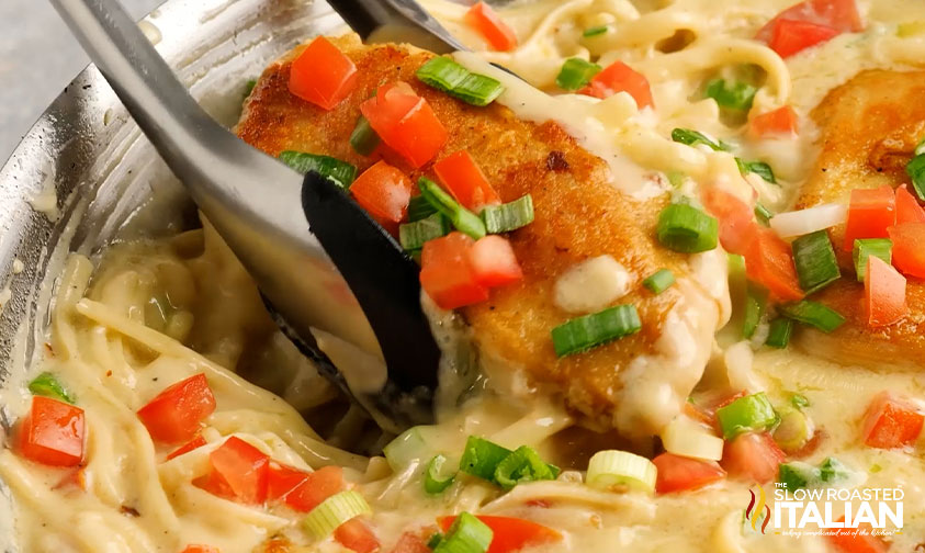 tongs lifting chicken out of skillet with tomatoes, green onion, pasta, and sauce