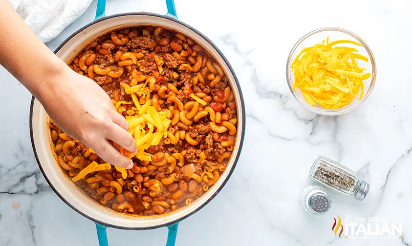 sprinkling shredded cheese over pot of chili mac