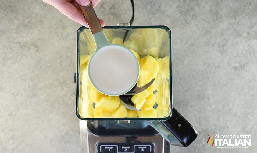 holding measuring cup of coconut milk over blender with pineapple