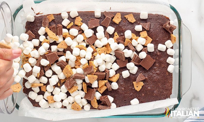 sprinkling marshmallow, graham cracker pieces, and chocolate over pan of brownies