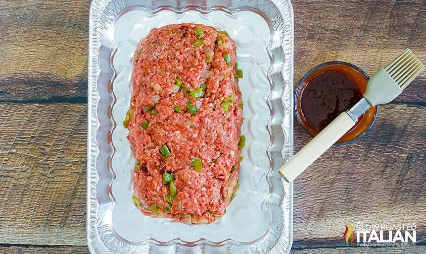 uncooked meatloaf in foil pan next to bowl of bbq sauce and basting brush