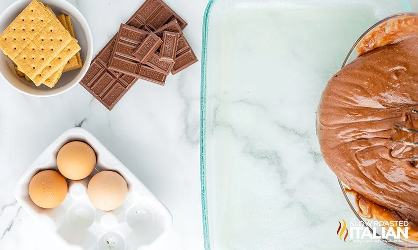 pouring chocolate cake batter into glass pan with eggs, grahams, and chocolate nearby