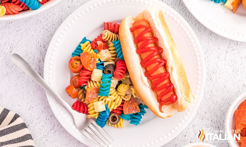 plate with tri color pasta salad and a hot dog in a bun with ketchup