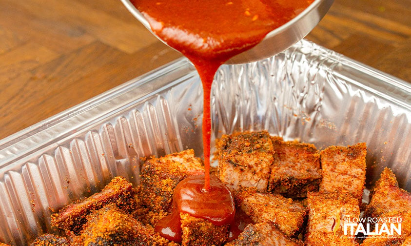 pouring barbecue sauce over seasoned burnt ends in foil pan