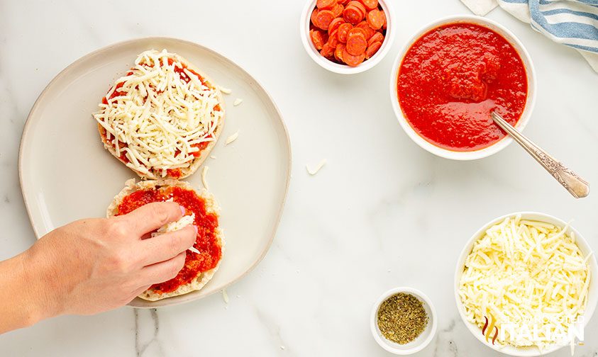 sprinkling shredded cheese on english muffin with pizza sauce