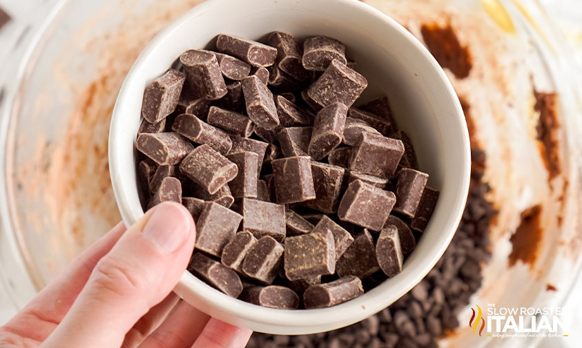 holding bowl of chocolate chunks over larger bowl with dough