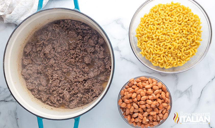 overhead: a large pot full of cooked ground beef, a large bowl full of uncooked macaroni, and a bowl of beans
