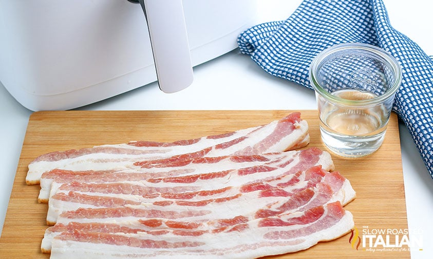 strips of raw bacon on cutting board with glass of water