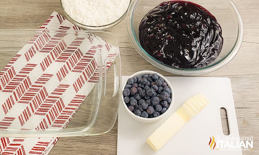 glass baking dish next to cake mix, butter, blueberries, and pie filling