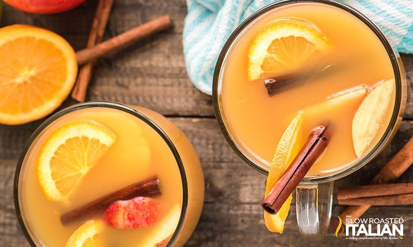 overhead: two clear mugs of hot cider with fruit and cinnamon sticks