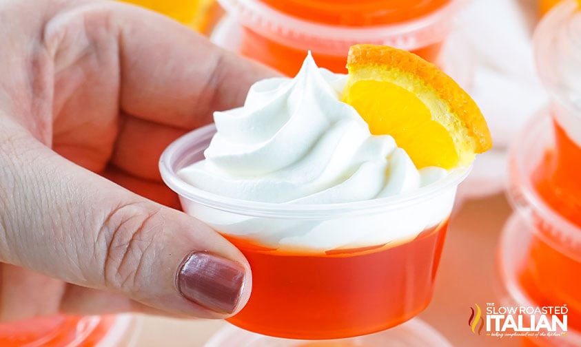 close up: holding orange jello shot with whipped cream and orange slice