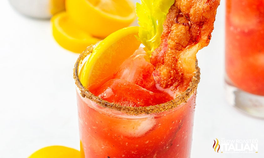 close up: glass of bloody mary cocktail rimmed in celery salt and garnished with bacon, celery, and lemon