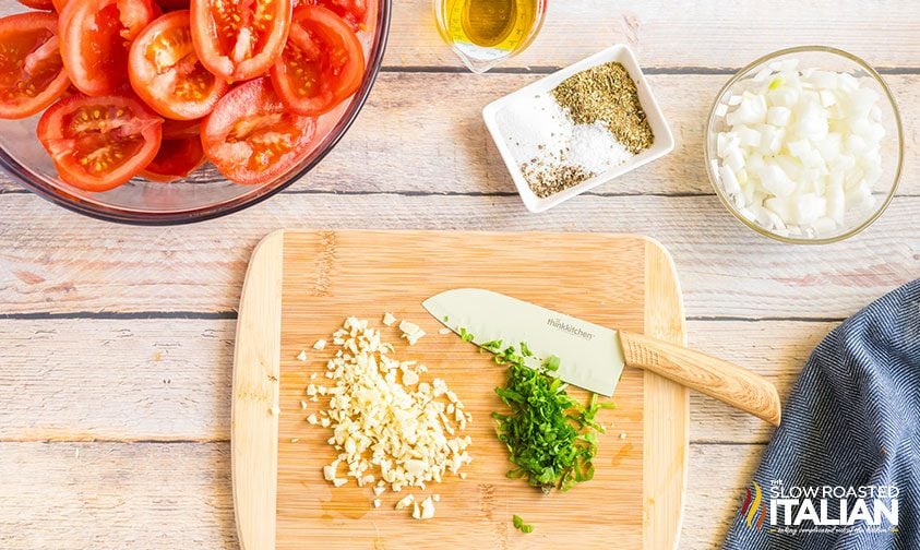 chopped garlic and herbs on cutting board next to bowls of tomatoes, onions, and spices