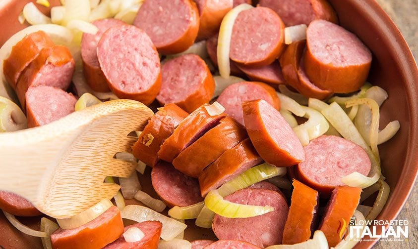 stirring sliced sausage and onions in skillet with wooden spoon