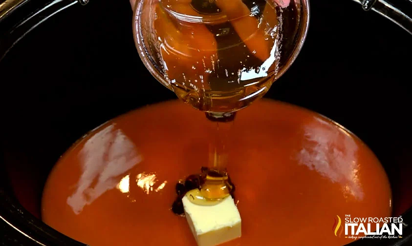 pouring honey into slow cooker with buffalo sauce and butter