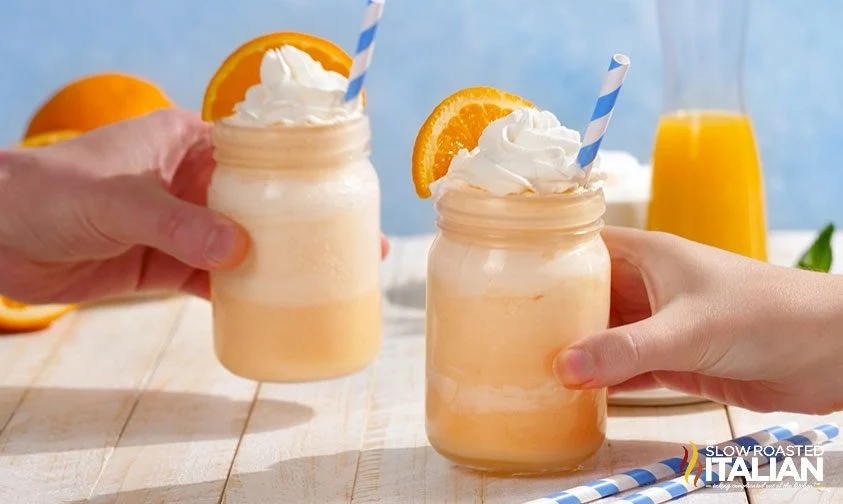 two people's hands, each holding an orange milkshake topped with whipped cream and orange slice in a mason jar with a straw