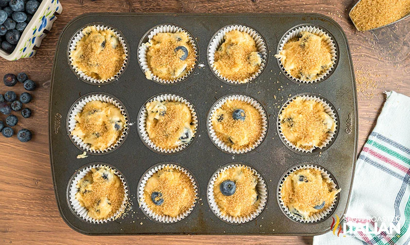 muffin pan with cupcake liners filled with blueberry batter topped with sugar crystals