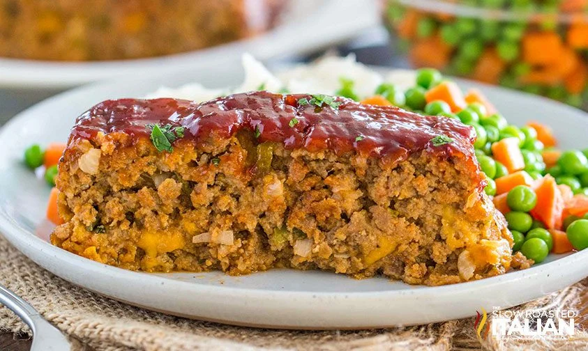 close up: slice of Cracker Barrel meatloaf topped with glaze, on a plate with mixed vegetables