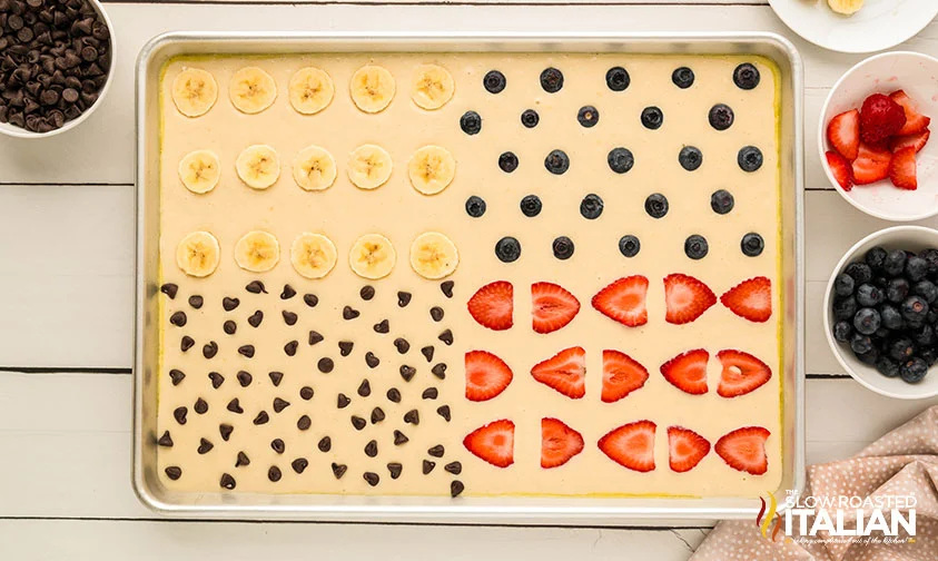 sheet pan pancake batter with 4 topping quadrants: banana slices, blueberries, chocolate chips, and sliced strawberries