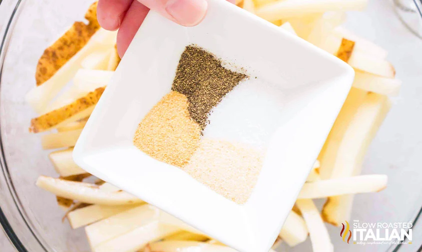 holding small bowl of seasonings over bowl of sliced potatoes for fries