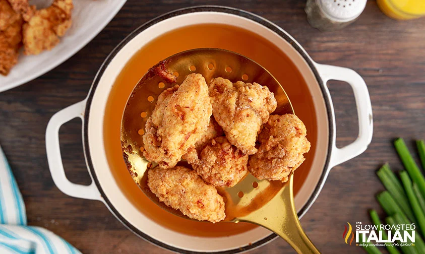 fried chicken pieces in strainer spoon held over pot of oil
