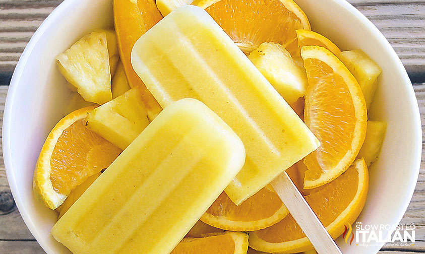 2 pineapple orange popsicles in a bowl of fruit