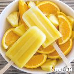 2 orange and pineapple popsicles in a bowl with sliced oranges and chunks of pineapple