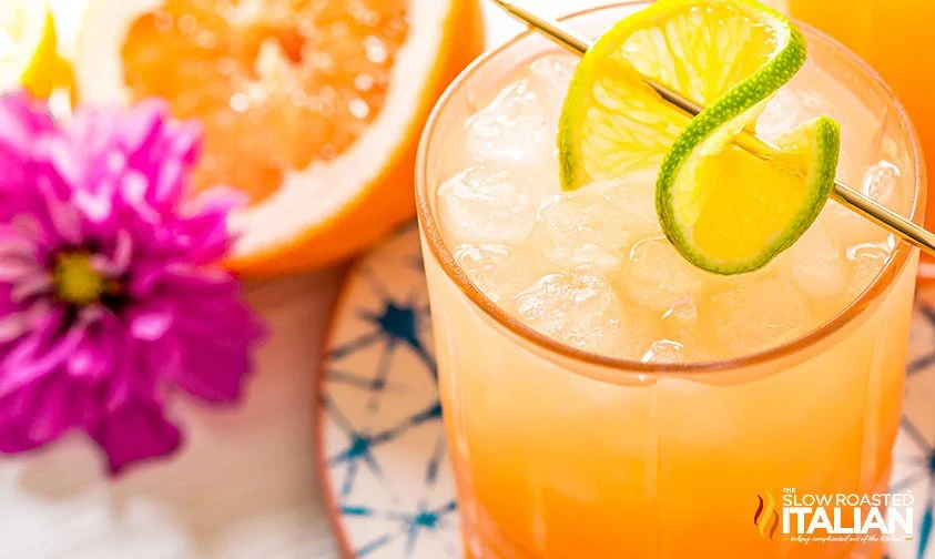 overhead: greyhound cocktail garnished with twist of lime, next to grapefruit half and purple flower