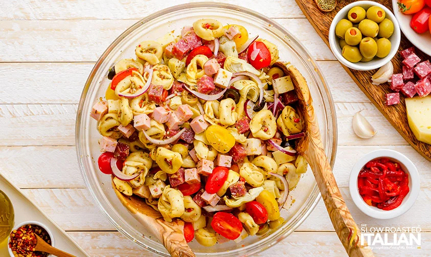 antipasto pasta salad tossed with dressing, in a bowl with wooden salad spoons