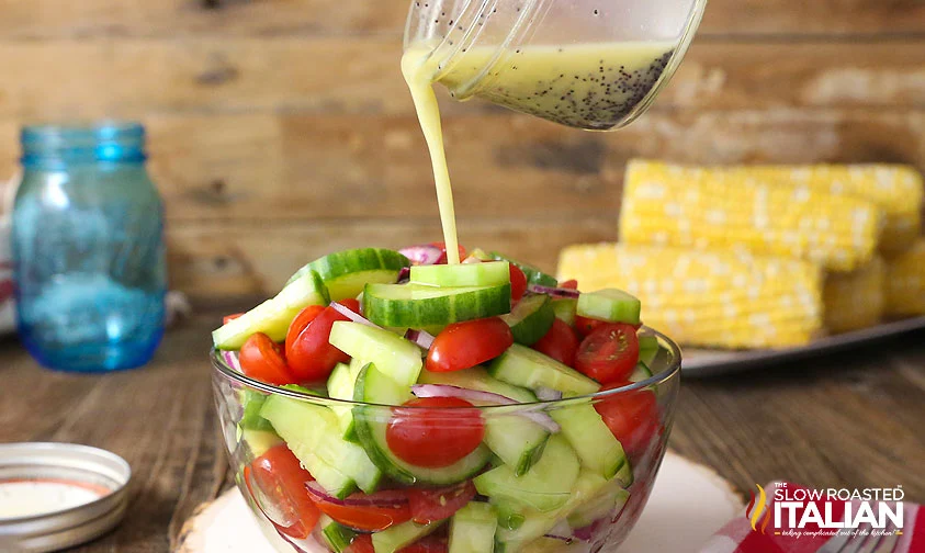 pouring lemon poppyseed dressing from a jar over bowl of tomato cucumber salad