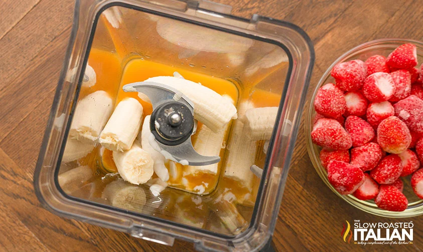 orange juice, bananas, and yogurt in blender next to bowl of frozen strawberries