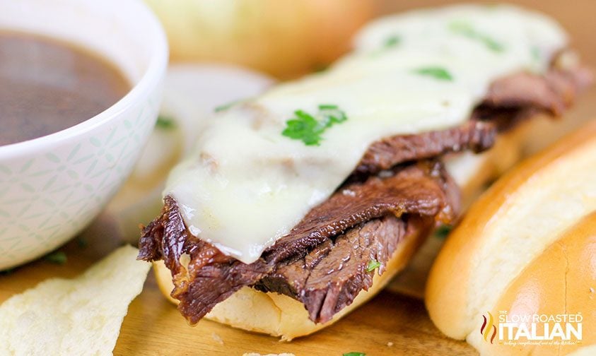 close up of cheese melted over beef on a hoagie roll next to a bowl of au jus