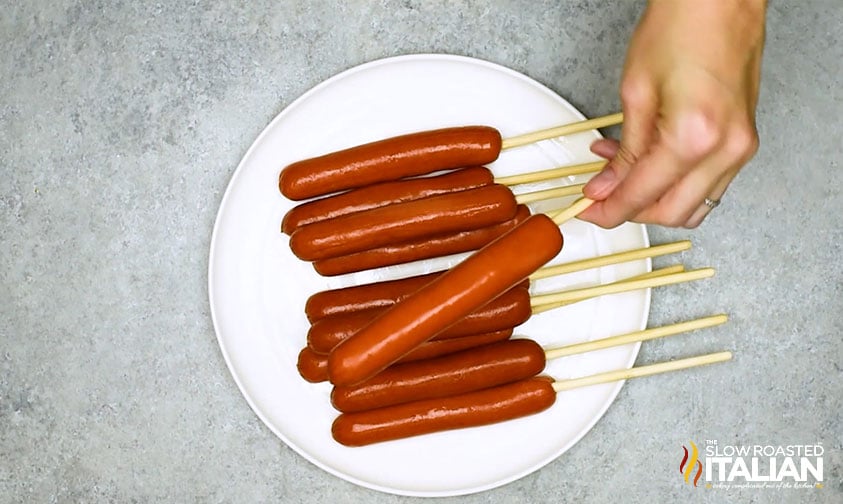 holding a skewered hot dog over a plate of others