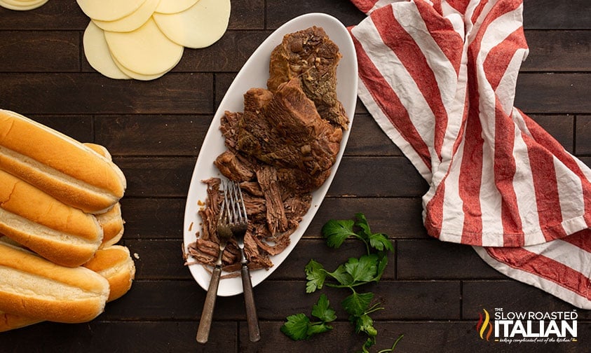 platter of slow cooked chuck roast surrounded by cheese slices, sandwich rolls, and a red and white striped towel