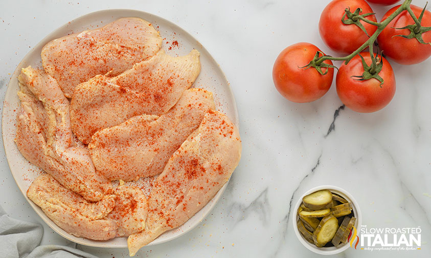 plate of seasoned, raw chicken next to tomatoes on the vine and bowl of pickle chips